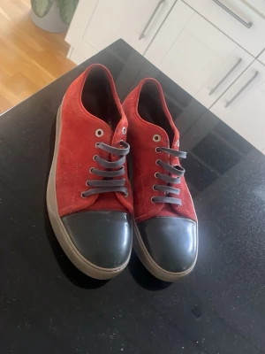 Röda sneakers med grå tå och snörning - Snygga sneakers i röd mocka med grå gummitå och grå snören. Skorna har en klassisk låg profil och silverfärgade öljetter. Yttersulan är beige och ger en schysst kontrast till det röda. Perfekta för dig som vill sticka ut lite extra.
