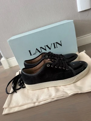 Svarta Lanvin sneakers i mocka och lack - Säljer ett par svarta Lanvin sneakers med ovandel i mocka och glansig lackdetalj på tån. Skorna har vita sulor och svarta skosnören. Klassisk låg modell med stilren design och metallöljetter. Perfekta för dig som gillar exklusiva och trendiga sneakers. Skorna är i väldigt gott skick och enbart använda ett fåtal gånger! Strl UK 8, passar 42.5-44. Kontakta för frågor/bilder🤩🤩OBS dustbag, box & snören tillkommer!