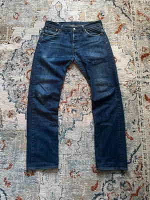 Levis 501 - Säljer ett par mörkblå jeans med rak passform och klassisk femficksdesign. Jeansen har en snygg tvätt och detaljer i metall vid fickorna. Passar perfekt till en avslappnad stil och är tillverkade i denim.