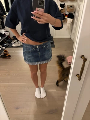 Blå jeanskjol med mönstrad kant - Säljer en blå jeanskjol i kort modell med en söt vitmönstrad kant längst ner. Kjolen har klassiska fickor fram och bak samt knapp och dragkedja framtill. Perfekt för dig som gillar en avslappnad och trendig stil.