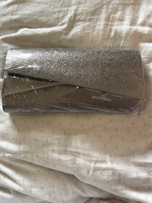 Silverfärgad glittrig kuvertväska - Snygg kuvertväska i silver med glittrig yta och elegant metallisk detalj på framsidan. Väskan har en stilren, rektangulär form och passar perfekt till att lyfta din outfit. Materialet är troligtvis syntet med glitterfinish.