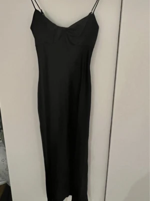Svart långklänning från Gina Tricot - Säljer en svart långklänning från Gina Tricot med tunna axelband och en smickrande, figurnära passform. Klänningen har en enkel och stilren design i glansigt material som ger en lyxig känsla. Perfekt för dig som gillar minimalistisk stil.