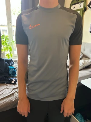 Grå sporttröja från Nike - Snygg grå sporttröja från Nike med korta ärmar och orange logga på bröstet. Tröjan har mörkare grå partier på ärmar och sidor, och är tillverkad i ett lätt och stretchigt funktionsmaterial som andas. Perfekt för träning eller fotboll.