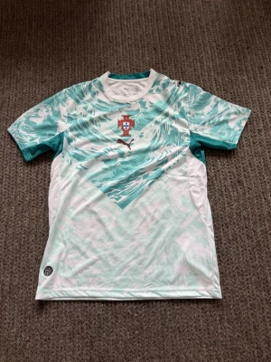Portugal fotbollströja Puma grön/vit - Snygg Portugal fotbollströja från Puma i vitt och mintgrönt med vågigt mönster. Tröjan har korta ärmar, rund hals och Portugal-emblem på bröstet. Perfekt för träning eller match!