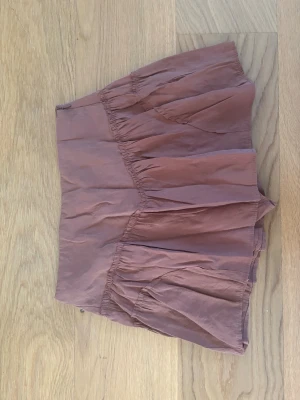 Brun volangkjol i bomull - Kort brun kjol med volangdetaljer och avslappnad passform. Kjolen är gjord i ett lätt bomullstyg och har en mjuk, rynkad design som ger en luftig känsla. Den har insydda shorts