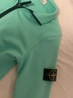 Turkos halfzip hoodie Stone Island - Snygg turkos hoodie från Stone Island med halv dragkedja framtill och klassisk logopatch på ärmen. Tillverkad i mjukt bomullsmaterial och har ribbade muddar vid ärmslut och nederkant. Perfekt för en chill och stilren look.