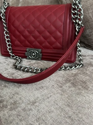 Vinröd Bag axelväska - Snygg vinröd   axelväska i quiltat läder med silverfärgad kedja och ikoniskt -spänne framtill. Klassisk boxig form och exklusiv känsla, perfekt för dig som vill sticka ut med en statementväska. Se bilden under väskan. Det finns små fläckar, men det är inget som syns när man bär väskan.  Väska har samma funktion 😁