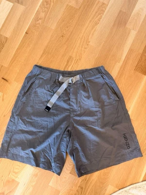 Grå shorts med bälte från Kenzo - Snygga grå shorts från Kenzo med justerbart bälte i midjan och två sidofickor fram. Shortsen har en avslappnad passform och är tillverkade i ett lätt, skrynkligt material som passar perfekt till sommaren. Kenzo-loggan syns diskret på ena benet.