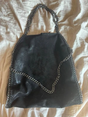 Svart handväska med kedjedetaljer - Snygg svart handväska i mjukt material med silverfärgade kedjor som handtag och längs kanterna. Väskan har en slouchy form och ger en cool, trendig vibe. Perfekt för dig som gillar statement-accessoarer och vill ha plats för allt du behöver. ( finns lite skavanker som du ser på bilderna )