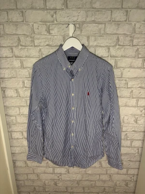 Ralph Lauren skjorta - Ralph Lauren skjorta i tunn och luftig bomull med klassiska blåvita ränder och röd broderad logga. En stilren och lättburen skjorta som passar perfekt under vår, sommar och varmare dagar. I mycket bra skick och väl omhändertagen. Hör av dig vid intresse 🙌