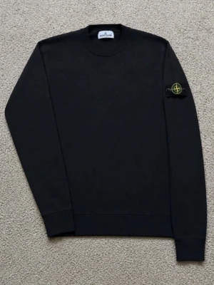 Svart sweatshirt från Stone Island - Snygg svart sweatshirt från Stone Island med klassisk rund halsringning och ikonisk patch med logga på vänster ärm. Tröjan har ribbade muddar vid ärmslut och nederkant. Perfekt för en clean och stilren look.