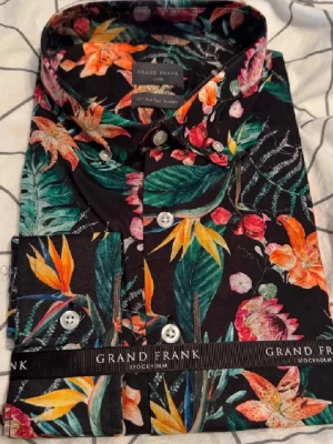 Blommig skjorta från Grand Frank - Säljer en färgstark blommig skjorta från Grand Frank med svart botten och mönster i grönt, orange, rosa och gult. Skjortan har klassisk krage, knappar framtill och långa ärmar. Perfekt för dig som vill sticka ut med din stil. Aldrig använd. Slim fit.
