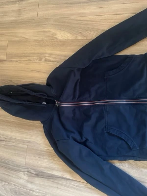 Mörkblå hoodie med dragkedja Moncler - Snygg mörkblå hoodie från Moncler med hel dragkedja och huva. Tröjan har fickor framtill och en Moncler-logga på ärmen. Dragkedjan har en röd, vit och blå detalj som ger en sportig touch. Perfekt för en avslappnad och trendig stil. Storlek S passar 170 ungefär. 