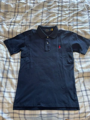 Mörkblå pikétröja från Polo Ralph Lauren - Säljer en klassisk och stilren pikétröja från Polo Ralph Lauren i marinblått med den ikoniska röda loggan på bröstet. Modellen är Custom Slim Fit, vilket ger en modern och mycket snygg passform.  Detaljer:  * Märke: Polo Ralph Lauren * Storlek: M (Custom Slim Fit) * Färg: Marinblå med röd broderad logga * Axelbredd: 50 cm * Längd: 69 cm * Skick: I absolut nyskick! Endast använd 3 gånger och mycket väl omhändertagen. Inga blekningar, hål eller fläckar.                  Skickar inom 2 dagar! 📦 