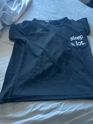 Svart pyjamaströja från SHEIN - Svart kortärmad pyjamaströja från SHEIN med tryck på bröstfickan där det står 'sleep a lot.' i vitt. Mjuk och bekväm, perfekt för slappa dagar eller nätter. Klassisk rund halsringning och enkel design.
