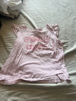 Ljusrosa topp med text från Lindex - Söt ljusrosa topp från Lindex med trycket 'Sprinkled Dreams' framtill och små färgglada detaljer. Toppen har korta ärmar med volang och är gjord i mjuk bomull, perfekt för en chill och lekfull stil.