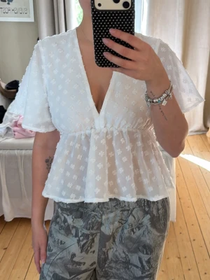 Vit blus med volang och v-ringning - Söt vit blus med v-ringning och volang nedtill. Blusen har korta ärmar och ett diskret blommigt mönster i tyget. Perfekt för dig som gillar romantiska detaljer och en luftig känsla.