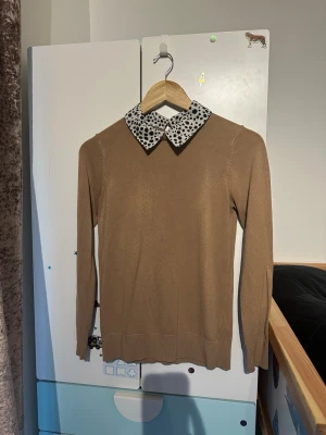 Beige stickad tröja Dorothy Perkins - Snygg beige stickad tröja från Dorothy Perkins med lång ärm och en vit krage med svartprickigt mönster. Tröjan har en klassisk, smal passform och är perfekt för dig som gillar en stilren look med en twist.