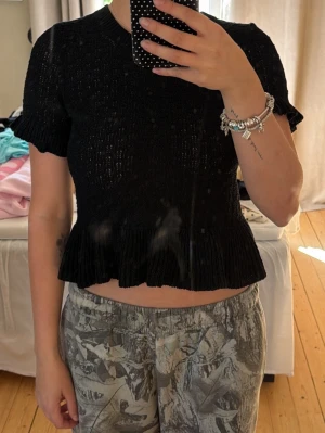 Svart croppad topp med volang - Säljer en svart croppad topp med korta ärmar och volangkant nedtill. Toppen är stickad och har en lätt ribbad struktur som ger en snygg detalj. Perfekt att styla med både jeans och kjol.
