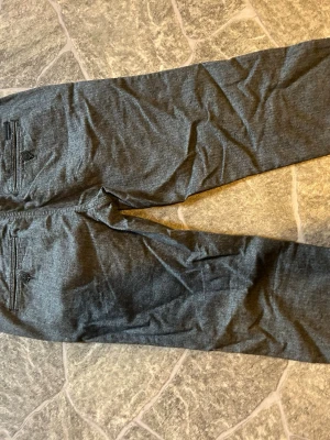 Grå kostymbyxor från Jack & Jones - Snygga grå kostymbyxor från Jack & Jones i klassisk rak modell. Byxorna har två bakfickor med knapp och diskret fiskbensmönster i tyget. Perfekta för en stilren look och passar till skjorta eller t-shirt.