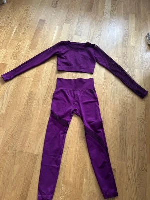 Lila träningstights och crop top set - Snyggt matchande träningsset i lila med långärmad crop top och höga tights. Setet har meshdetaljer på ena ärmen och benen för extra ventilation och stil. Perfekt för gymmet eller yoga. Materialet är stretchigt och följsamt för maximal rörelsefrihet.