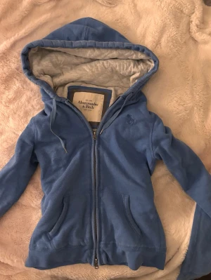 Blå hoodie vintage Abercrombie & Fitch - Blå zip-hoodie från Abercrombie & Fitch med mjukt grått foder i huvan. Klassisk design med dragkedja, två fickor fram och dragsko i huvan. Perfekt för chill dagar och enkel att matcha med jeans eller joggers.