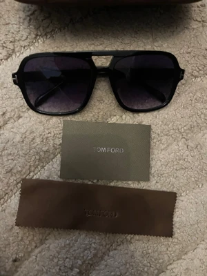 Svarta Tom Ford solglasögon - Snygga svarta solglasögon från Tom Ford med markerad båge och mörka glas. Modellen har en modern, kantig form och kommer med originalfodral och putsduk. Perfekta för dig som vill ha en stilren accessoar med exklusiv känsla.