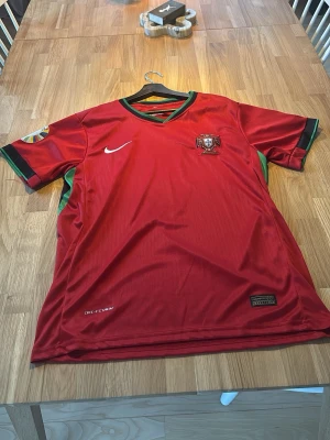 Portugal fotbollströja Ronaldo #7 - Säljer en röd Portugal fotbollströja från Nike med gröna detaljer och officiellt landslagsmärke på bröstet. På ryggen står det 'Ronaldo' och nummer 7. Tillverkad i lätt och ventilerande material, perfekt för match eller träning.