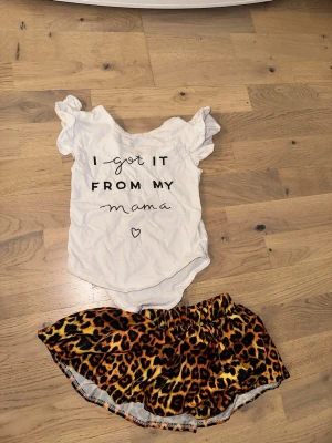 Babyset med textbody och leopardkjol - Sött babyset med vit body med texten 'I got it from my mama' och volangärmar. Tillhörande kjol i mjukt tyg med leopardmönster i brunt, svart och gult. Perfekt för en liten fashionista som gillar att sticka ut.Babyset med textbody och leopardkjol
