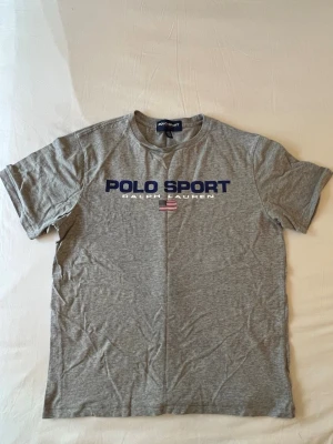 Grå Polo Sport t-shirt från Ralph Lauren - Snygg grå t-shirt från Polo Sport Ralph Lauren med blå logga och amerikansk flagga på bröstet. Klassisk passform och rund halsringning. Perfekt till jeans eller shorts för en avslappnad look.