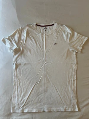 Vit t-shirt från Hollister - Klassisk vit t-shirt från Hollister med rund halsringning och liten broderad logga på bröstet. Tillverkad i mjuk bomull och har en enkel, stilren design som passar till det mesta. Perfekt basplagg för alla tillfällen.