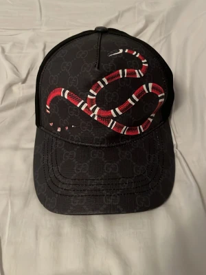 Svart Gucci keps med ormtryck - Snygg svart keps från Gucci med ikoniskt ormtryck i rött, vitt och svart på framsidan. Kepsen har diskret GG-mönster över hela ytan, mesh på baksidan och justerbart band med gröna och röda detaljer. Perfekt för dig som vill sticka ut med en lyxig accessoar.