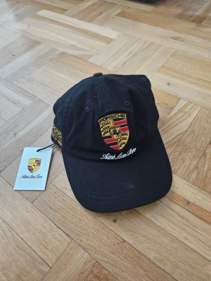 Svart Porsche keps Aimé Leon Dore - Svart keps med böjd skärm och broderad Porsche-logga framtill samt texten Aimé Leon Dore. Märket syns även på sidan. Kepsen har klassisk passform och justerbar rem bak. Materialet är troligtvis bomull.