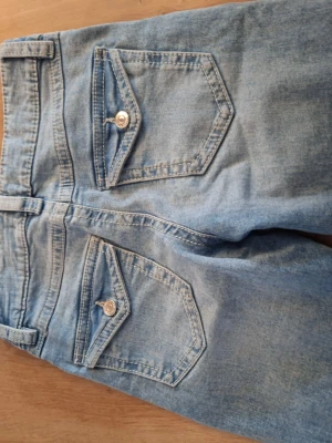 Ljusblå slitna jeans med ficklock - Säljer ett par ljusblå jeans med lite bootcut ben och slitningar på både lår och knä. Jeansen har två bakfickor med lock och knappdetaljer. Tillverkade i klassisk denim med en avslappnad vibe. Perfekta för dig som gillar en trendig och casual stil.