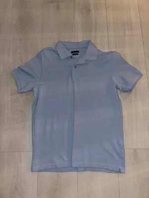 Ljusblå pikétröja från Massimo Dutti - Säljer en stilren ljusblå pikétröja från Massimo Dutti. Tröjan har klassisk krage, korta ärmar och knappar vid halsen. Tillverkad i mjuk bomull för en bekväm känsla och perfekt för en clean look.