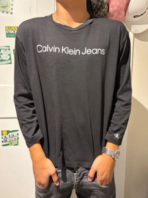 Svart långärmad topp Calvin Klein Jeans - Svart långärmad topp från Calvin Klein Jeans med vit logotyp framtill och liten CK-detalj på ärmen. Avslappnad passform och rund halsringning. Perfekt för en stilren och enkel look.