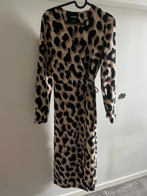 Leopardmönstrad omlottklänning Monki - Snygg långärmad omlottklänning från Monki med leopardmönster i beige och svart. Klänningen har knyt i midjan och v-ringad hals. Perfekt för dig som vill sticka ut med ett trendigt djurmönster.