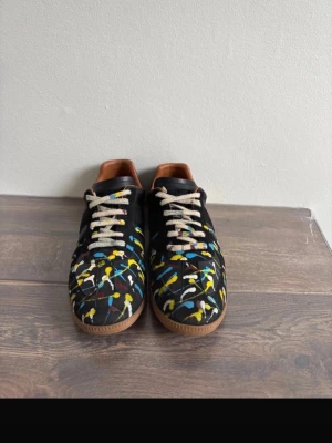 Maison Margiela svarta sneakers - Unika svarta sneakers från Maison Margiela med färgstänk i gult, blått och vitt över hela ovandelen. Skorna har beige snören, bruna gummisulor och insida i brunt läder. Materialet är en mix av läder och mocka för en cool kontrast.