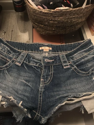 Blå jeansshorts med fransar - Säljer ett par blå jeansshorts med råa fransar i nederkant och vita kontrastsömmar. Shortsen har fem fickor, bälteshällor och klassisk knappgylf. Perfekta för sommaren och ger en avslappnad vibe. Dom har blivit för små för mig och vill att dom ska komma till användning för dom är i bra skick