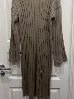 Beige ribbstickad lång klänning - Säljer en beige ribbstickad lång klänning med långa ärmar och slits framtill. Klänningen har en rak passform och är tillverkad i ett mjukt stickat material som känns skönt mot huden. Perfekt för lager-på-lager och enkel att styla med boots eller sneakers.