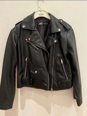 Svart skinnjacka från ZARA - Snygg svart skinnjacka från ZARA med klassisk bikerstil, silvriga dragkedjor och knappar. Jackan har axelklaffar, flera fickor med dragkedja och en cool asymmetrisk dragkedja framtill. Perfekt för en edgy look. Finns ett tillhörande bälte!