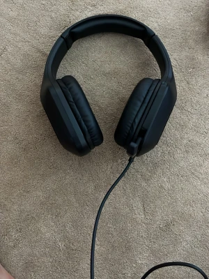 Svarta over-ear hörlurar med sladd - Säljer ett par svarta over-ear hörlurar med mjuka, vadderade kåpor och justerbar bygel. Hörlurarna har en lång sladd med 3,5 mm kontakt, perfekt för gaming eller musik. Klassisk design i plast och konstläder som sitter bekvämt runt öronen.