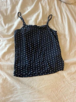 Mörkblå pyjamas med hjärtmönster - Säljer ett set med pyjamas i mjukt material, består av ett linne med smala axelband och ett par långa byxor. Båda delarna är mörkblå med små vita hjärtan över hela tyget. Perfekt för mysiga kvällar hemma. Sällan använd! 