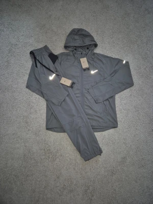 Nike Miller - Snyggt grått träningsset från Nike med jacka och byxor. Jackan har huva, dragkedja och reflekterande detaljer på bröstet och ärmarna. Byxorna har resår i midjan och reflekterande Nike-logga. Perfekt för löpning eller träning utomhus.