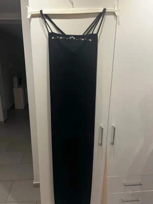 Svart långklänning med smala band - Elegant svart långklänning med smala axelband och dekorativ broderad detalj upptill. Klänningen har en rak och slimmad passform som ger en stilren siluett. Perfekt för dig som vill ha en enkel men snygg look.