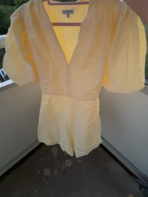 Gul kort jumpsuit med puffärmar - Superfin ljusgul jumpsuit med korta ben och djup v-ringning. Puffiga korta ärmar och knytband i midjan för en snygg siluett. Perfekt för varma dagar och ger en fräsch look. Materialet känns lätt och luftigt.