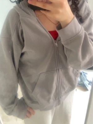 Grå zip hoodie med huva - Enkel och stilren grå hoodie med dragkedja framtill och stor ficka på magen. Hoodien har en klassisk huva och ribbade muddar vid ärmar och nederkant. Perfekt för chill dagar eller när du vill ha en avslappnad look.