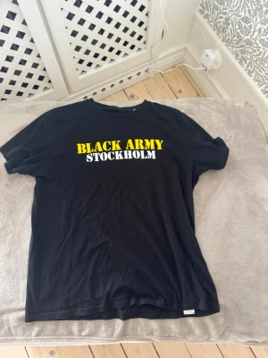 Aik t-shirt Black Army Stockholm tryck - AIK t-shirt med trycket 'BLACK ARMY STOCKHOLM' i gult och vitt på bröstet. Klassisk passform med korta ärmar och rund hals. Perfekt för dig som vill visa ditt stöd för Black Army och AIK . Tillverkad i mjukt bomullstyg.