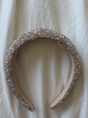 Glittrigt diadem med pärlor och stenar - Säljer ett brett diadem täckt av små pärlor och glittrande stenar i silver, vitt och roséguld. Diademet har en vadderad form som ger volym och är klätt i beige tyg på insidan för extra komfort. Perfekt för att ge outfiten en extra touch av glamour.