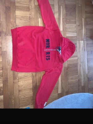 Röd hoodie med dragkedja från Morris - Snygg röd hoodie från Morris med halv dragkedja framtill och stor känguruficka. Huva med snörning och mörkblå text på bröstet. Perfekt för en avslappnad och sportig stil. Mjuk och skön bomullskänsla.